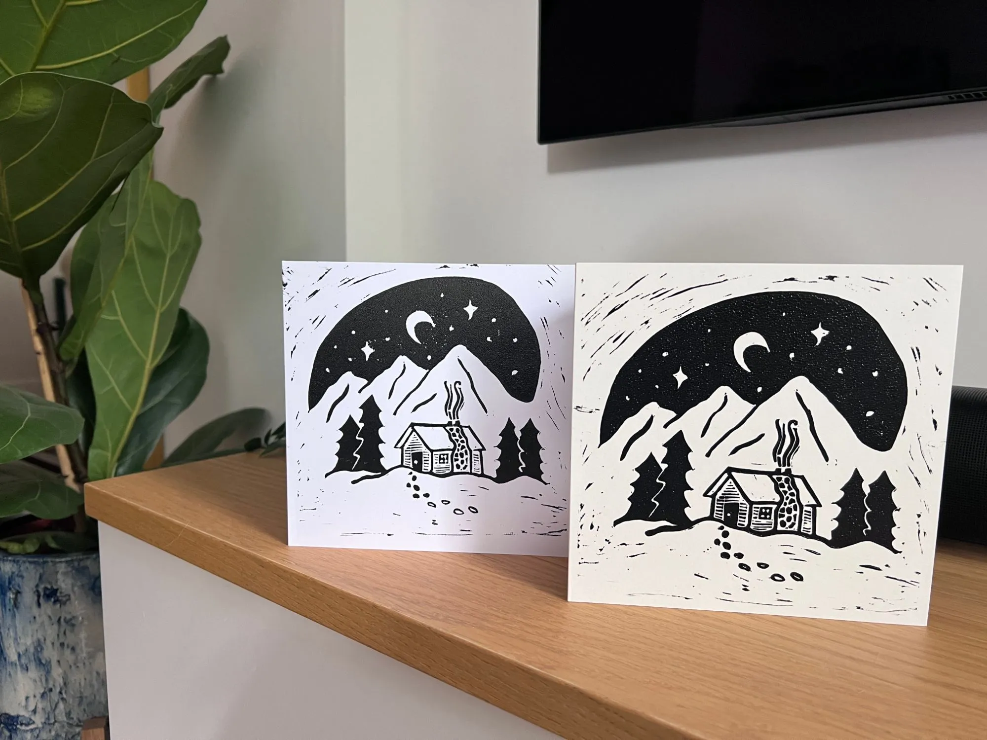 Two square lino-print Christmas cards of a cabin under a starry night sky, standing on a wooden shelf