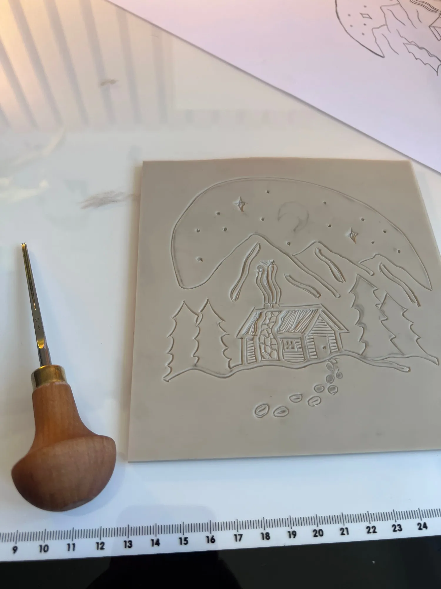 A carved lino block of a cabin scene beside a wooden-handled carving tool